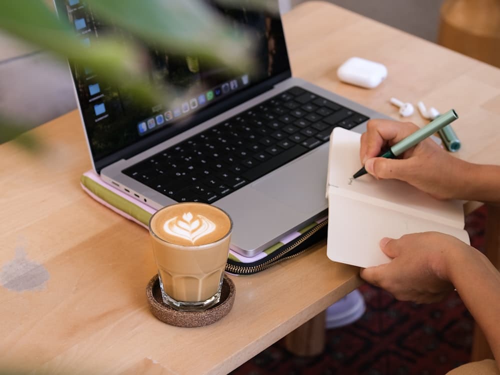 A writer drafting in a notebook beside a laptop and coffee